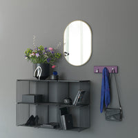 Black wire shelving with a purple hook, green and purple flowers, and a round gold-framed mirror on a gray wall.