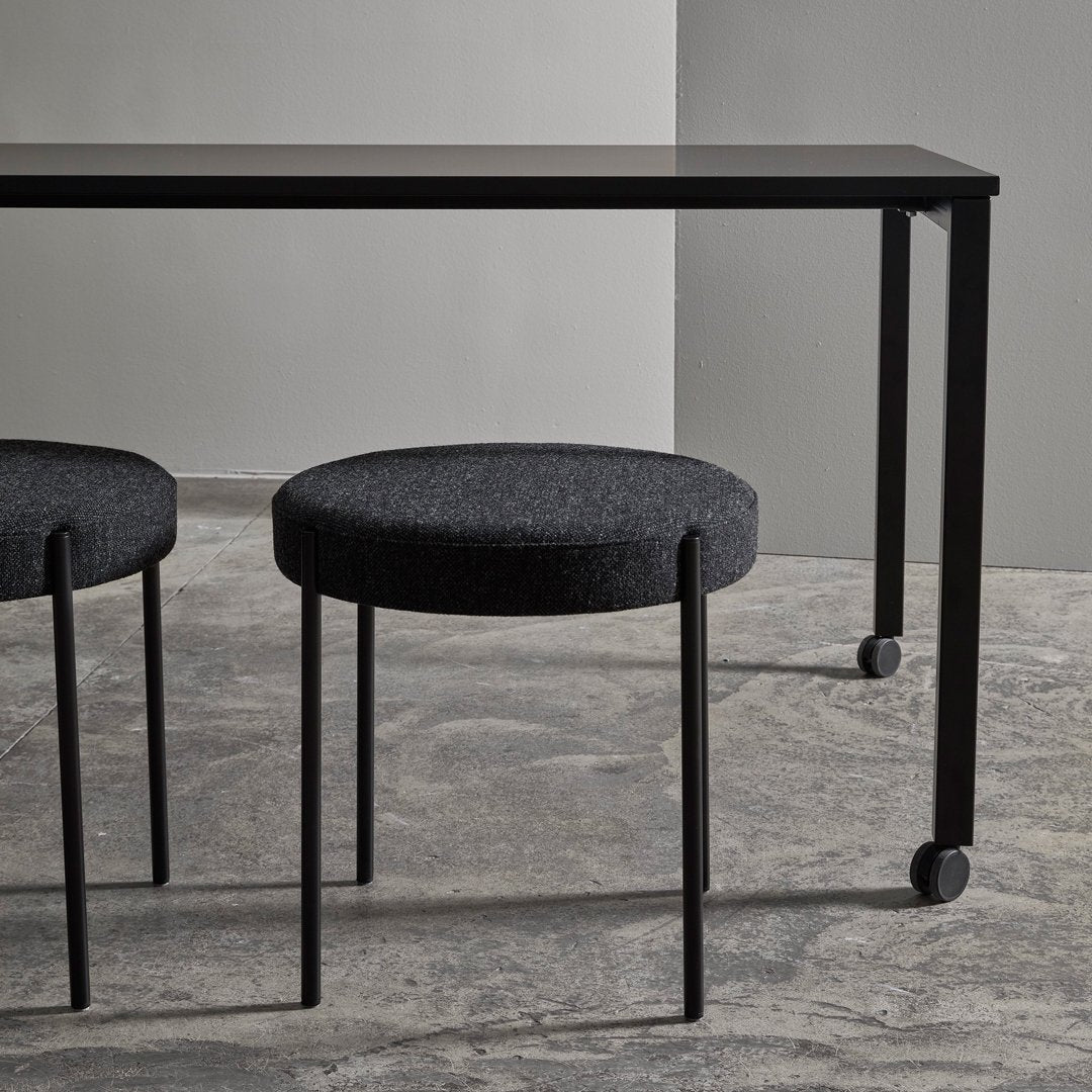 Black modern table with sleek design and two round black fabric stools on a textured gray floor.
