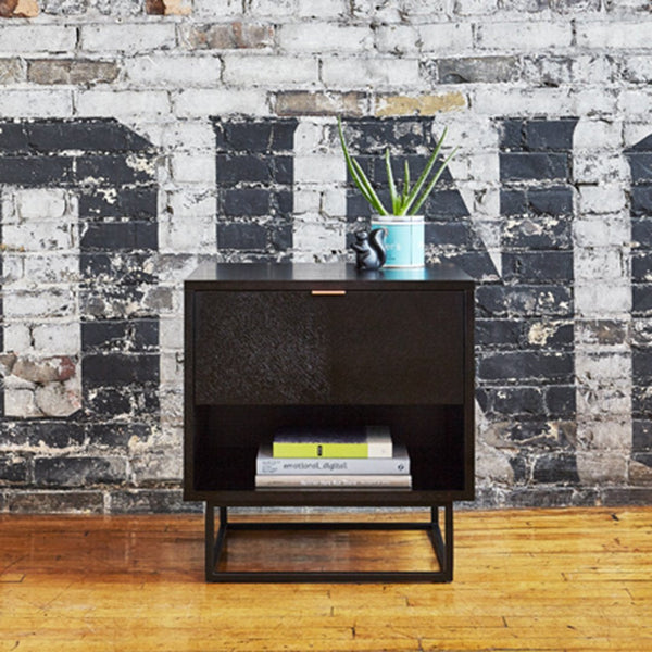 Black nightstand with a textured surface, featuring a sleek design against a rustic brick wall.