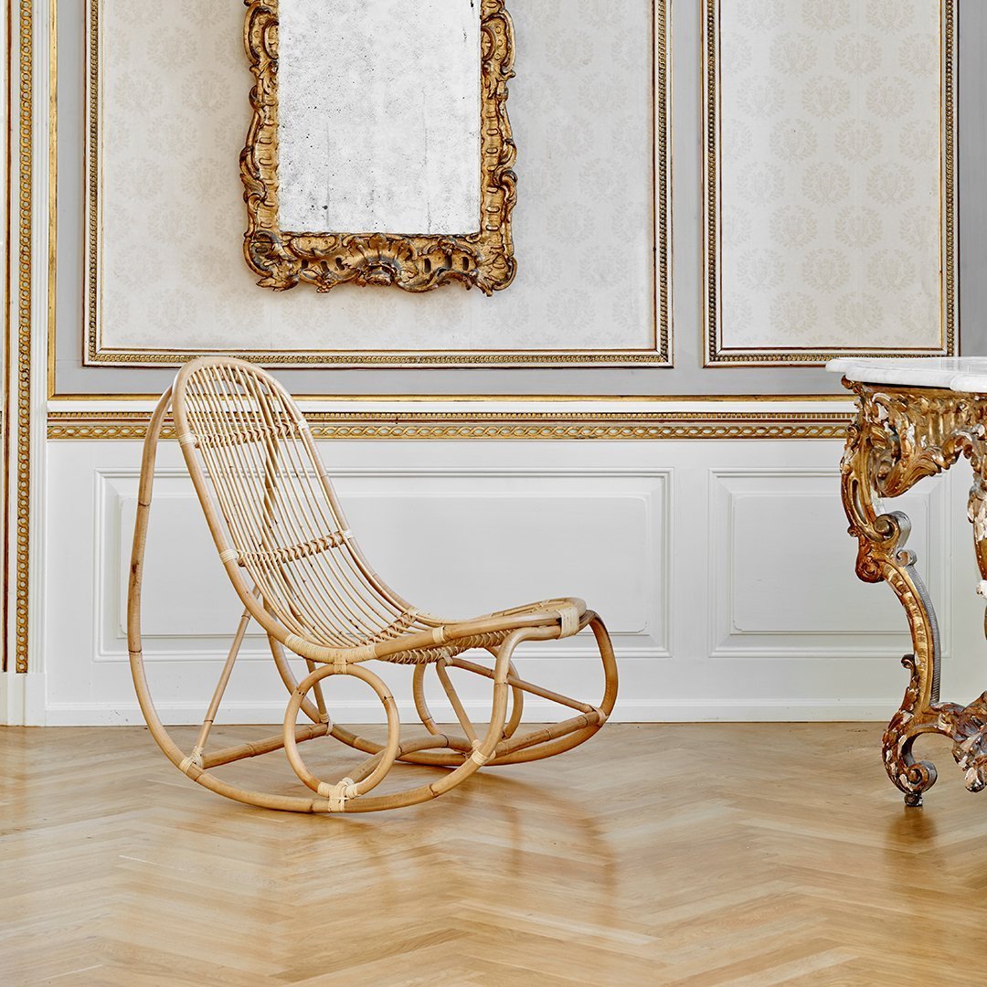 Natural rattan rocking chair in a light, airy room with white and gold accents, featuring elegant flooring and ornate details.