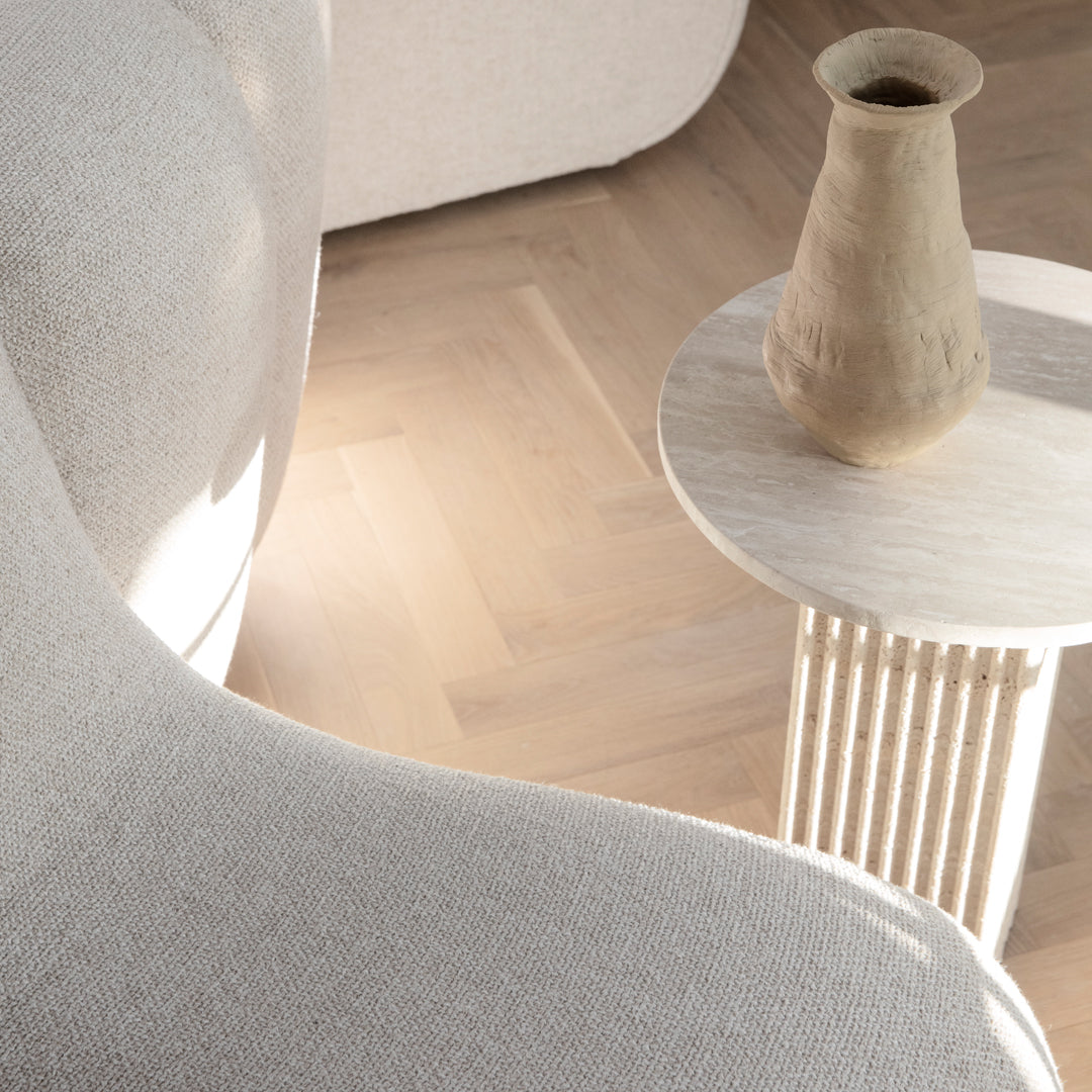 Neutral beige curved sofa with textured upholstery, light wood flooring, and a minimalist round side table in soft tones.