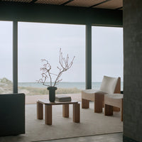 Minimalist living room featuring beige chairs, wooden furniture, and a black vase with branches on a light rug.