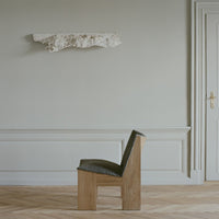 Green textured chair with natural wood legs against a soft gray wall and light herringbone floor. Modern minimalist design.