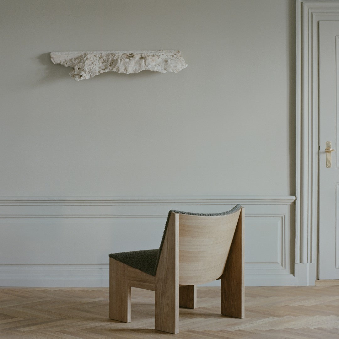 Natural wooden chair with a textured green fabric seat, against a soft gray backdrop and pale wooden flooring.