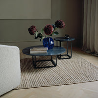 Round black coffee tables on a woven beige rug, accented by a vibrant blue vase and deep red flowers.