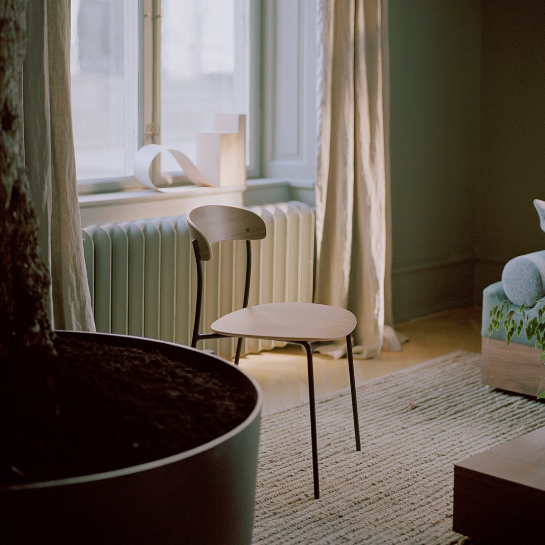 Minimalist wooden chair with a light beige finish, set against soft green walls and a textured beige rug.