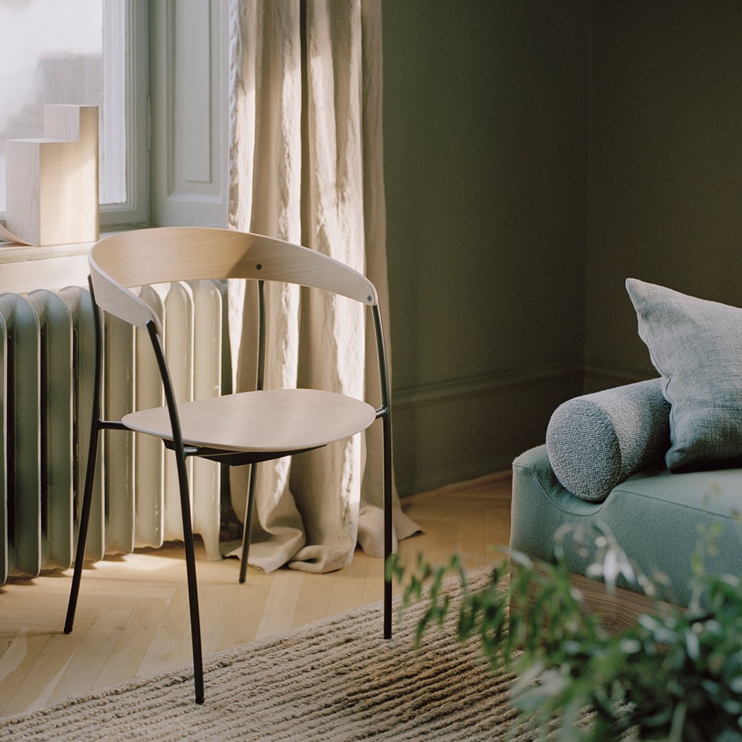 Modern chair with a natural wood seat, black metal legs, and a minimalist design, set in a stylish muted green room.