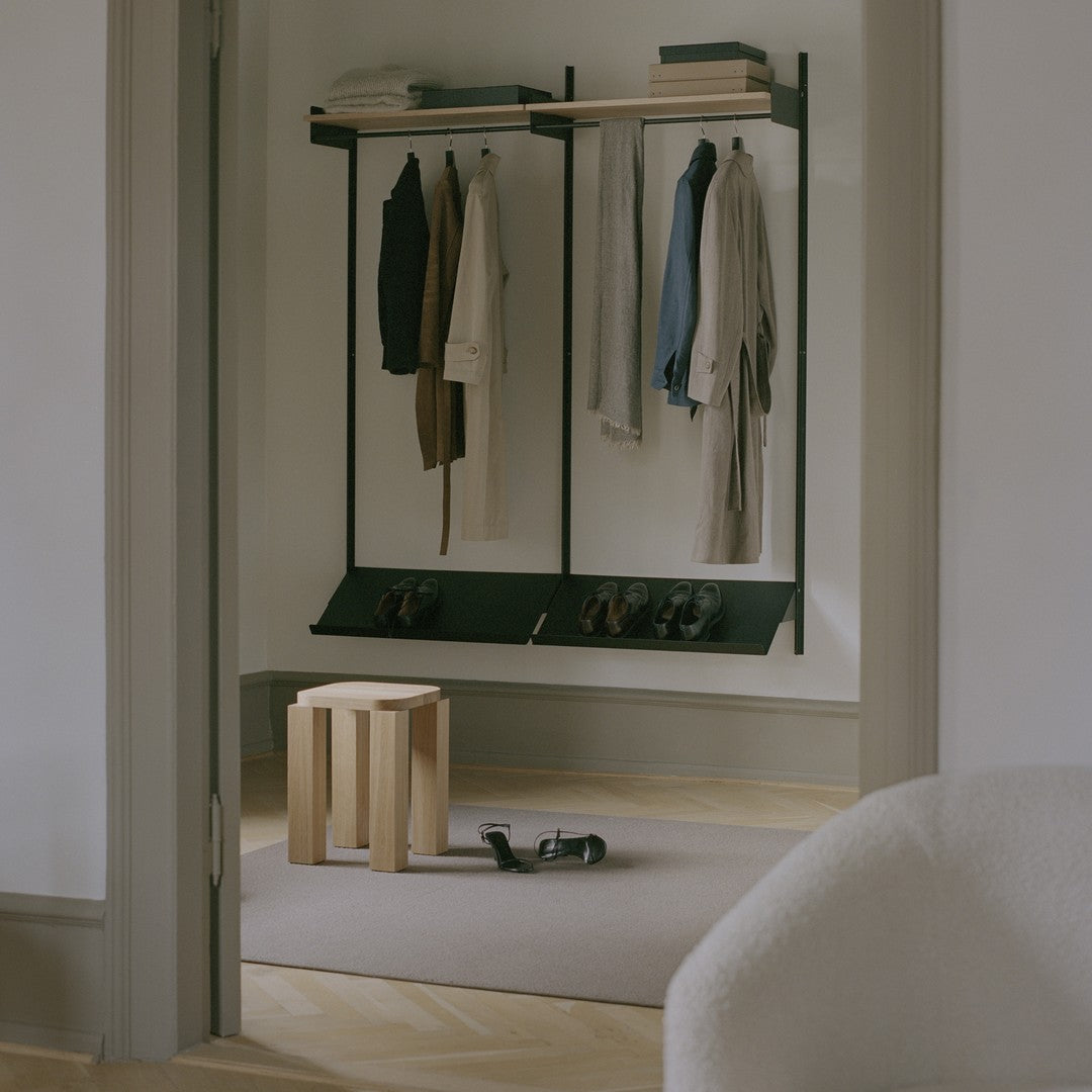 Minimalist entryway featuring black rack, neutral-hued clothing, wooden stool, and beige area rug.
