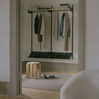 Minimalist entryway featuring black rack, neutral-hued clothing, wooden stool, and beige area rug.