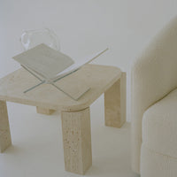 Beige textured coffee table with rounded edges, paired with a soft white fabric armchair and a clear vase.