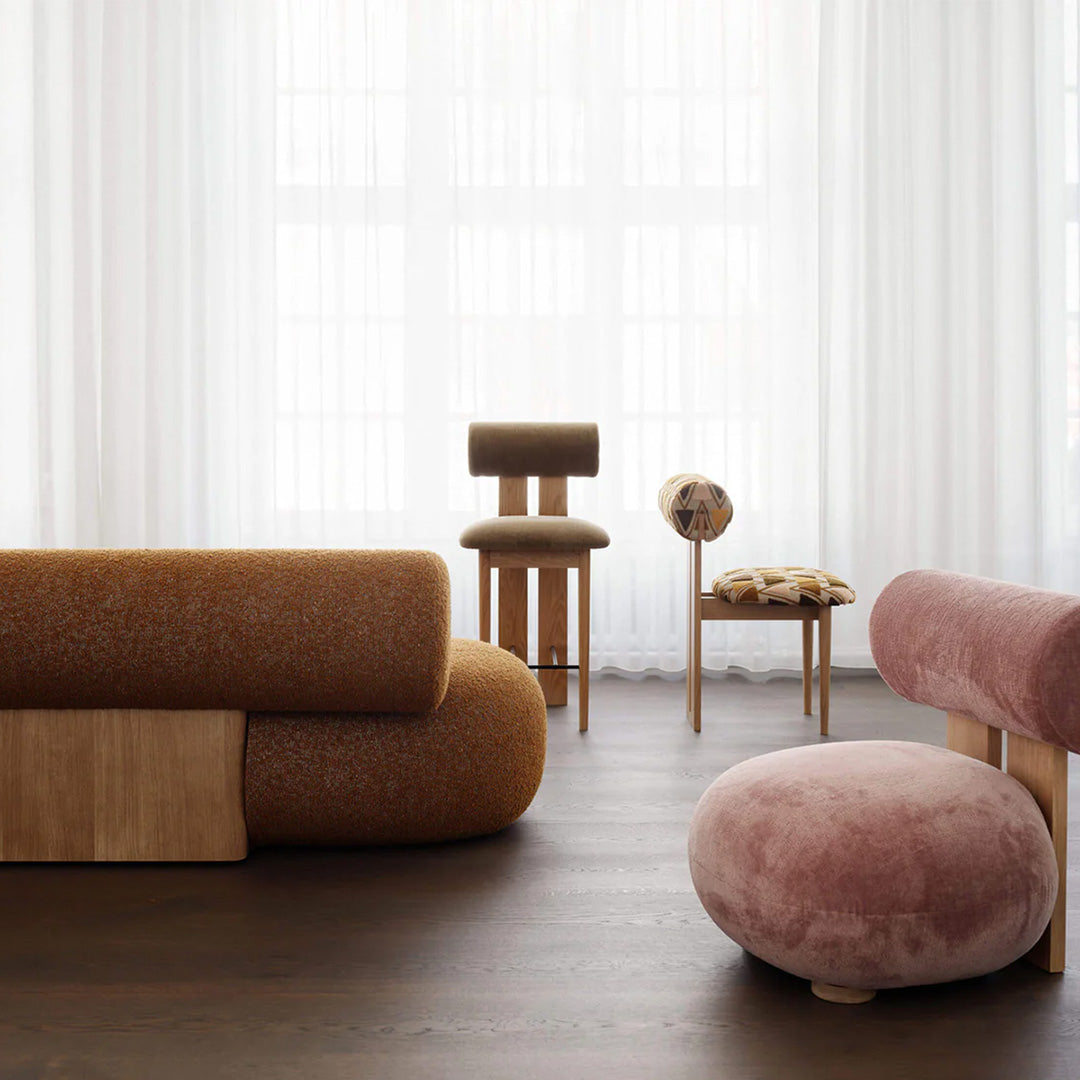 Sofa in warm orange fabric, pink circular stool, and patterned beige chair against a bright, airy backdrop.