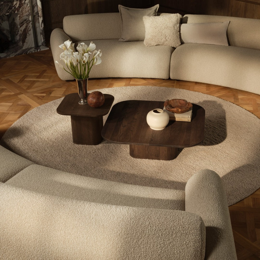 Beige curved sofa with textured fabric, paired with dark wood tables and a neutral round rug, accentuated by natural decor.