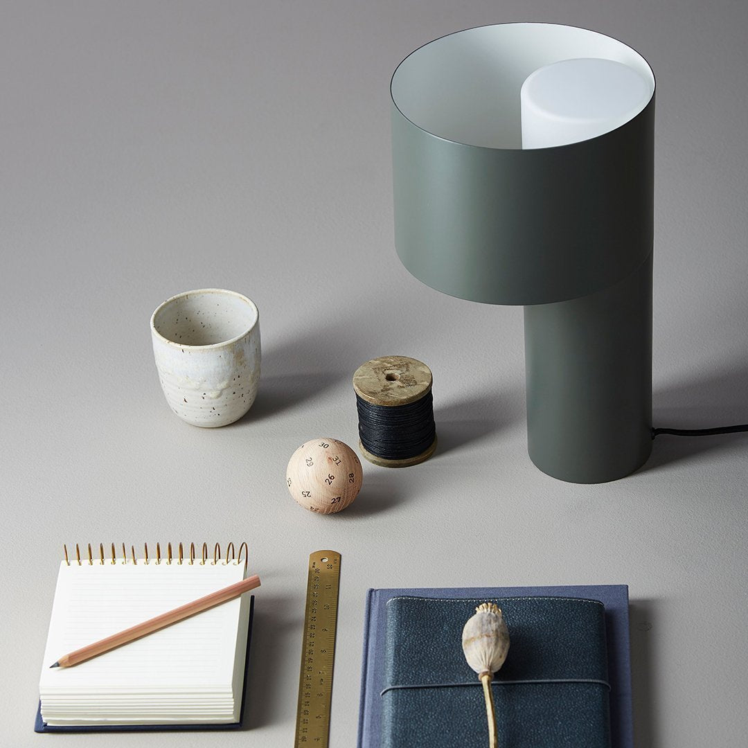 Modern green lamp with smooth matte finish, accompanied by textured ceramics and light wood accents on a neutral background.
