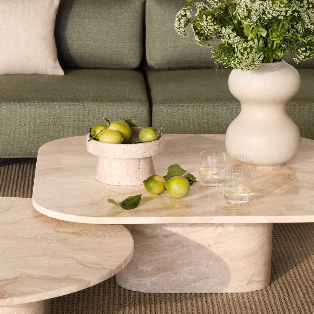Light beige marble coffee table with smooth curves, topped with a green fruit bowl, against a dark green sofa backdrop.