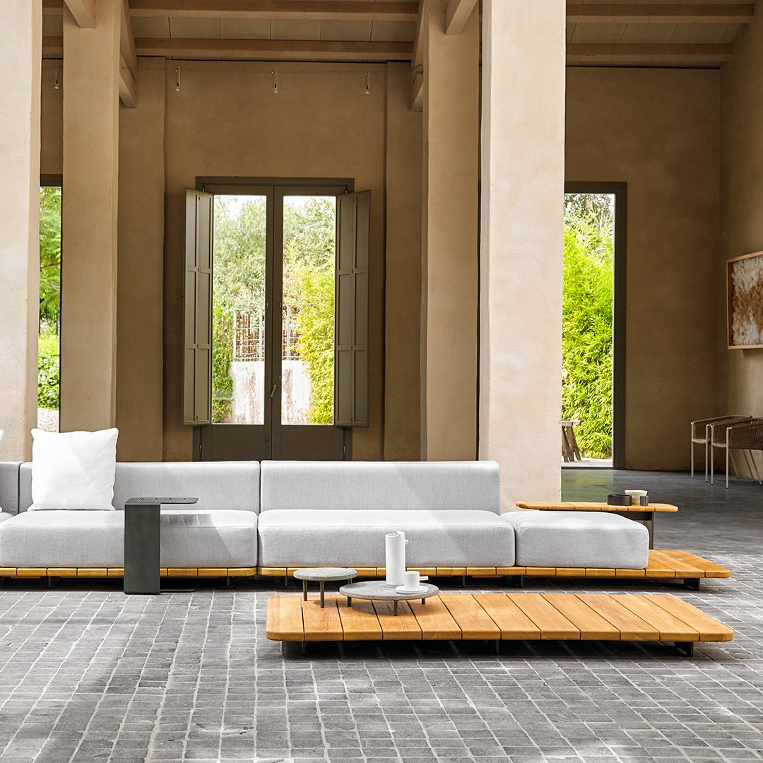 Modern gray sectional sofa with natural wood accents, set against a light earthy backdrop and patterned stone flooring.