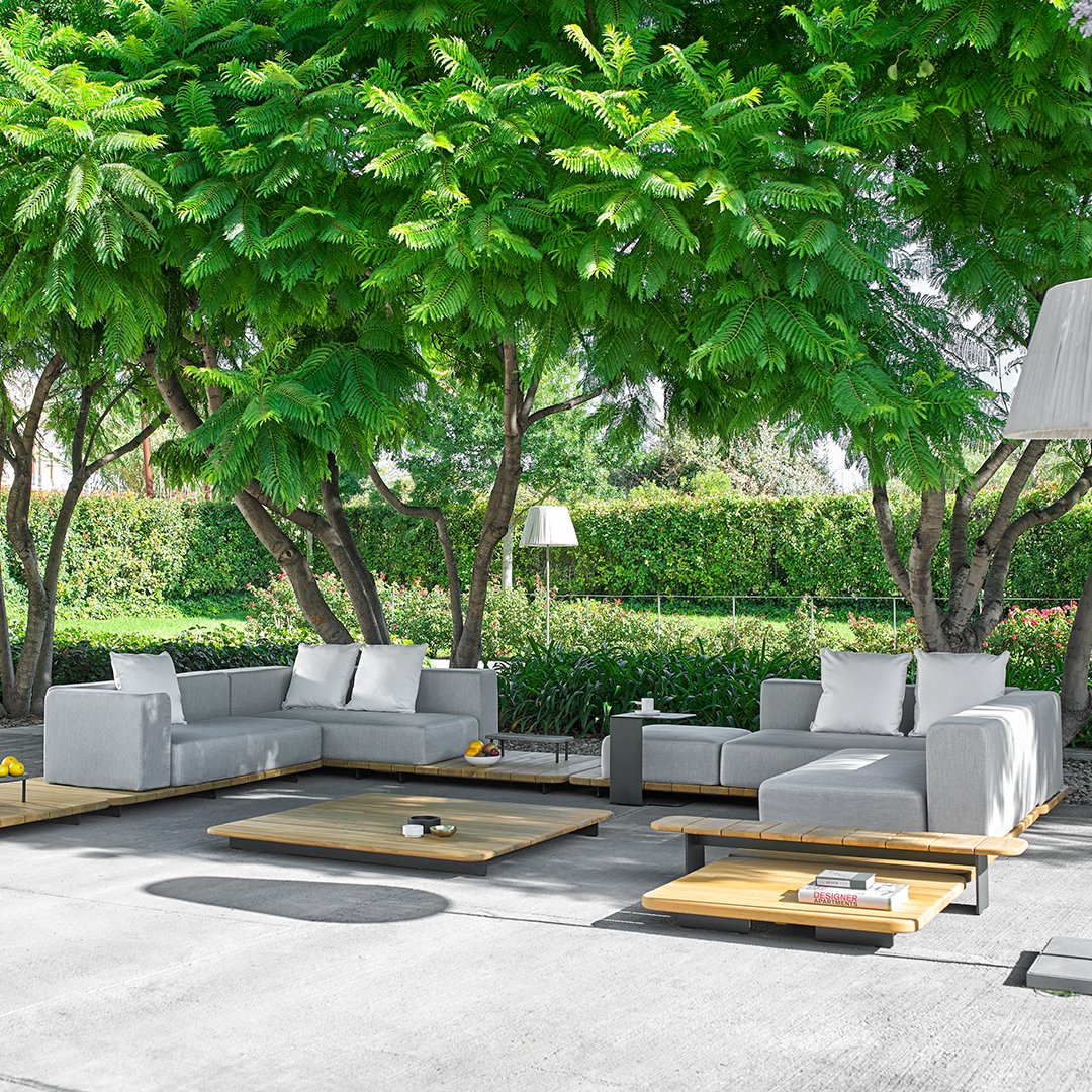Modern outdoor sectional sofa in light gray with natural wood accents, set against lush green foliage.