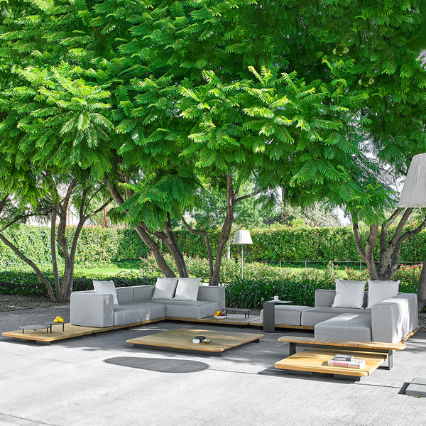 Gray sectional sofa with white cushions, set on a light wood platform under lush green trees. Modern outdoor design.
