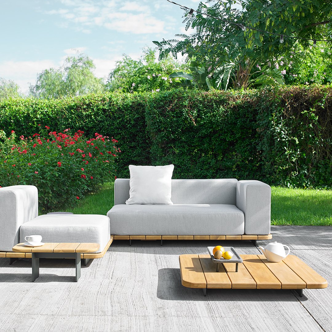 Grey outdoor sofa with wooden accents, surrounded by lush greenery and vibrant flowers, set on a light stone patio.