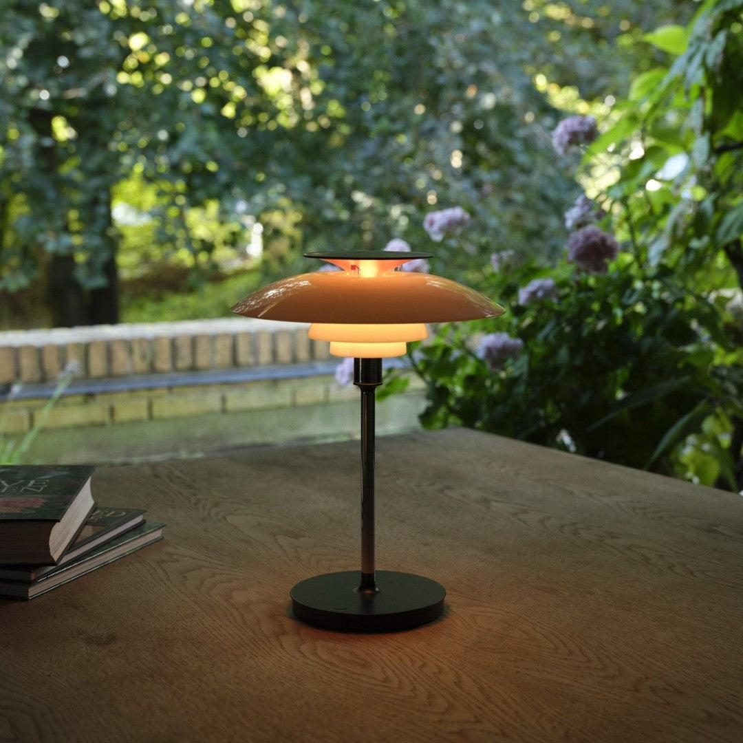 Modern table lamp with a sleek orange shade, glowing warm yellow light, and a black metal base, set against greenery.