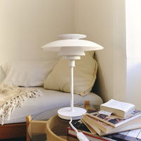 White modern table lamp with layered design atop a wooden side table, surrounded by neutral-toned books and cushions.