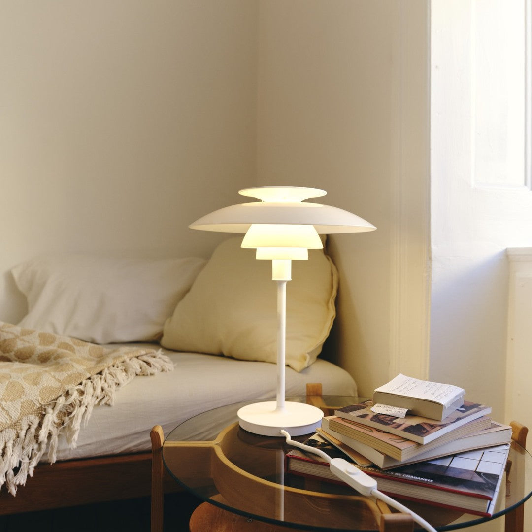 Modern white lamp with layered design, emitting warm yellow light, against a neutral beige and white bedroom backdrop.