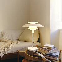 Modern white lamp with layered design, emitting warm yellow light, against a neutral beige and white bedroom backdrop.