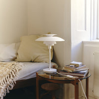 White table lamp beside a beige cushion on a bed, with a textured cream throw and a glass-top table.