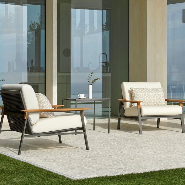 Beige and cream armchairs with wood accents, paired with a simple patterned throw pillow on a neutral outdoor rug.