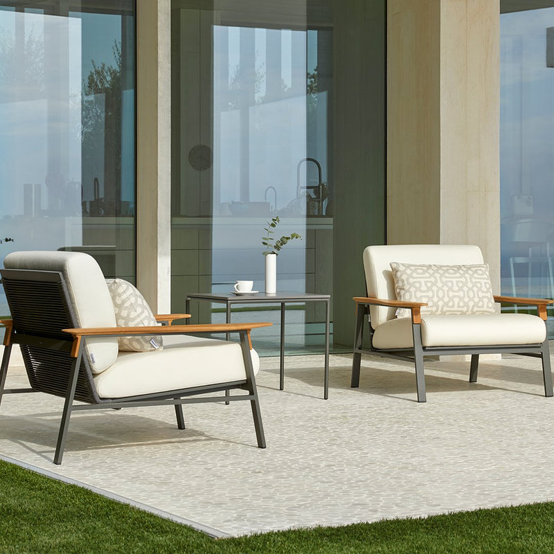 Beige and cream armchairs with wood accents, paired with a simple patterned throw pillow on a neutral outdoor rug.