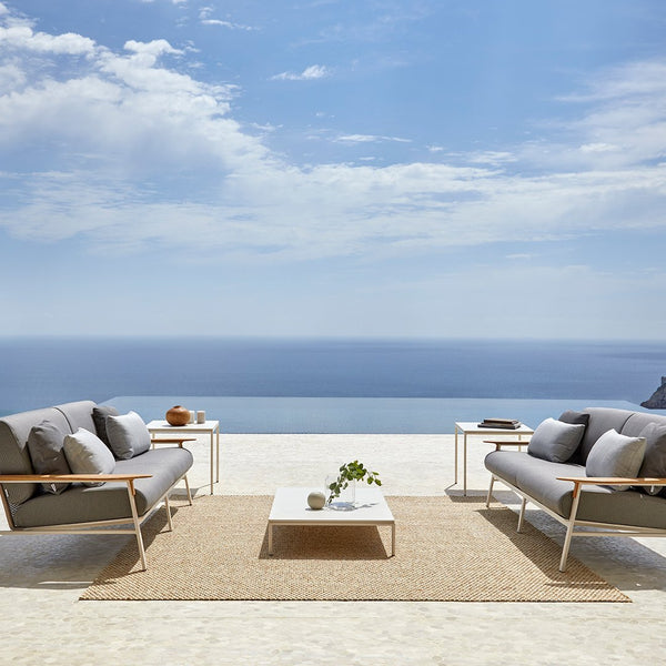 Gray outdoor sofas with white frames, paired with a beige rug and light-colored coffee table, against a blue ocean backdrop.