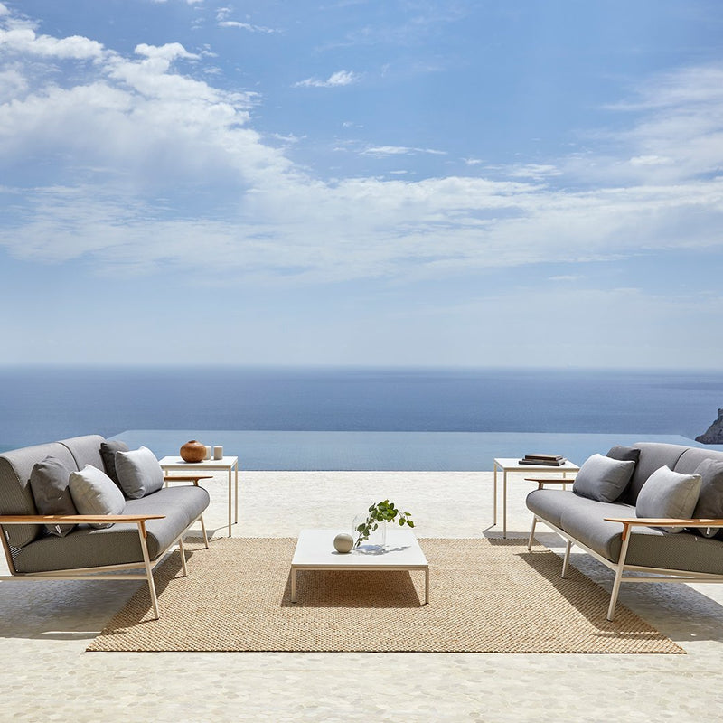 Gray outdoor sofas with white frames, paired with a beige rug and light-colored coffee table, against a blue ocean backdrop.