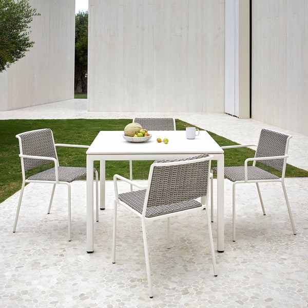 White outdoor dining set with gray woven seat cushions, set on a light stone patio surrounded by green grass.