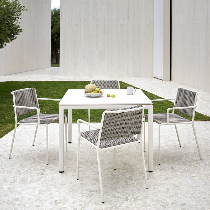 White outdoor dining set with gray woven seat cushions, set on a light stone patio surrounded by green grass.