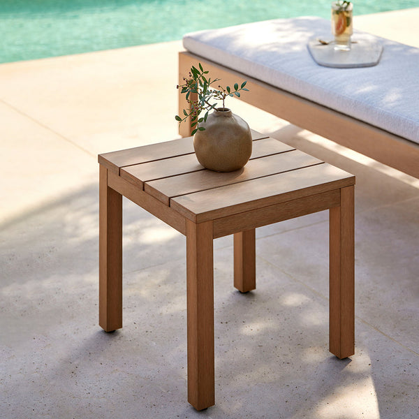 "Natural wood patio table in warm tones, featuring a slatted design, set beside a pool with soft beige and green accents."