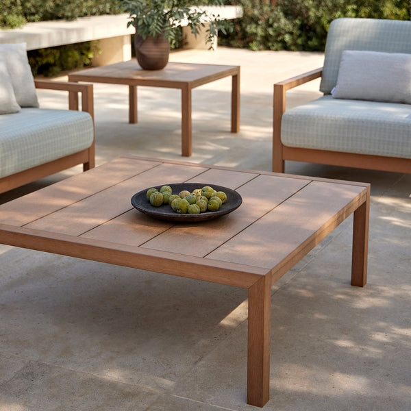Light wood patio furniture with soft blue cushions and a round black plate filled with green fruit on a stone surface.