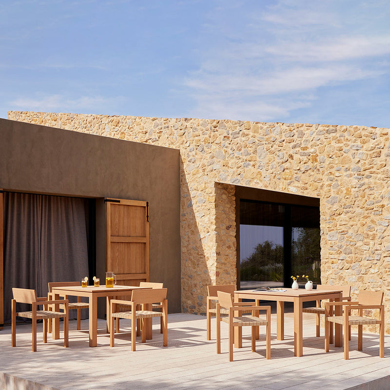 "Natural wood dining set with a sandy stone wall backdrop and a clear blue sky, perfect for outdoor settings."