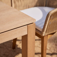 Light oak wooden table with a sleek finish, paired with a beige woven chair and white cushion for a modern look.