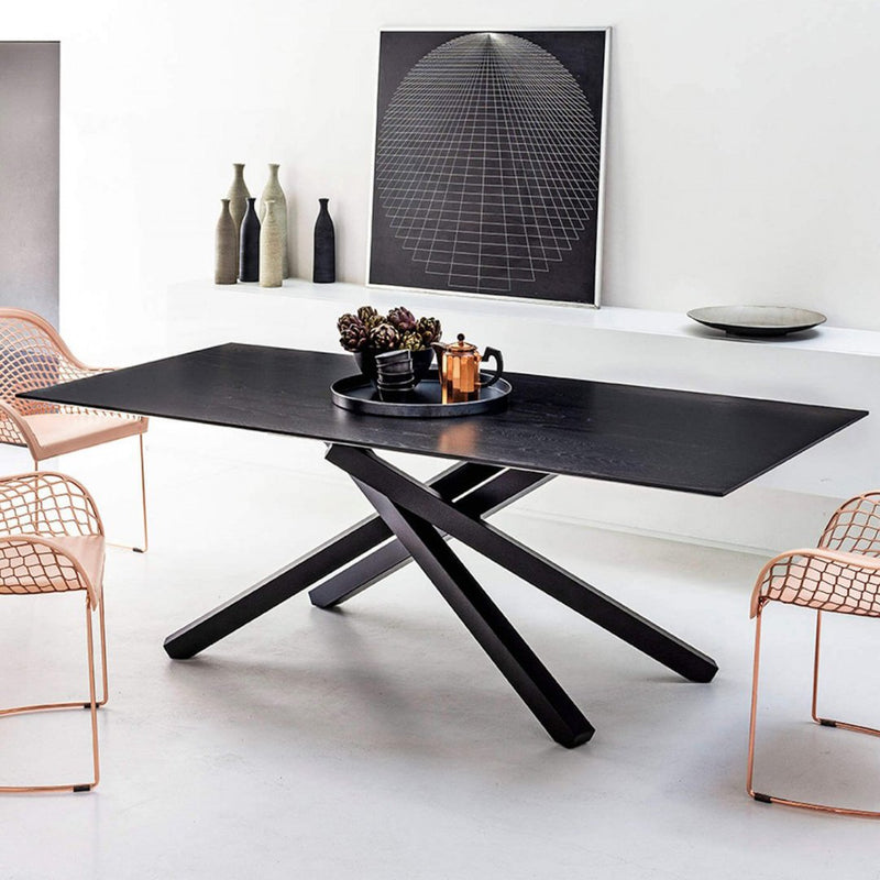 Stylish black dining table with geometric base, surrounded by modern rose gold chairs and minimalistic décor.