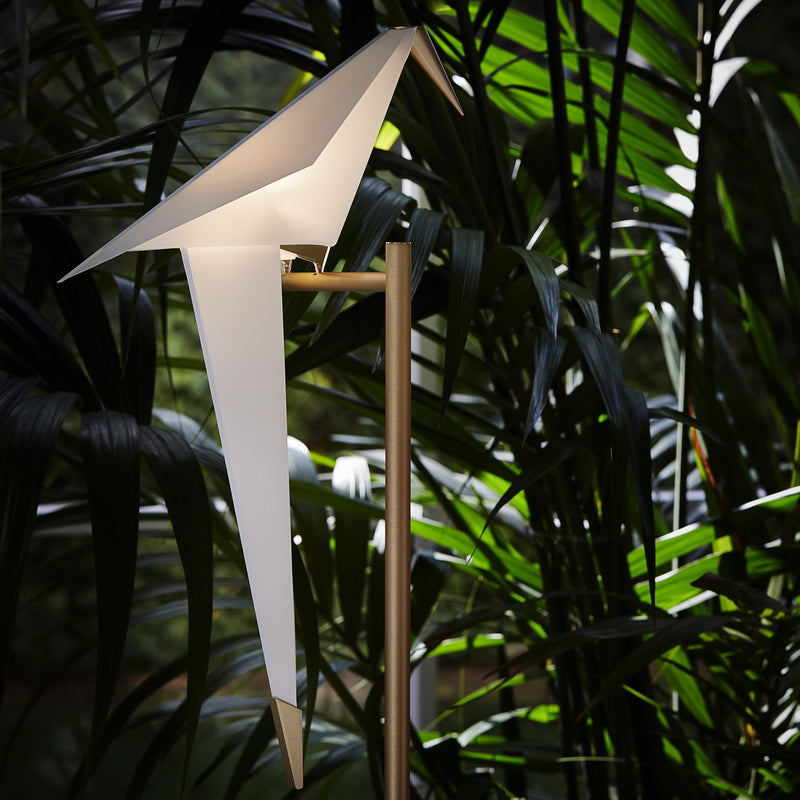 Modern white origami-style lamp with a sleek gold stand, surrounded by lush green tropical foliage.