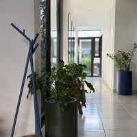 Blue coat rack and green cylindrical planter in a bright hallway with potted plants and neutral tile flooring.