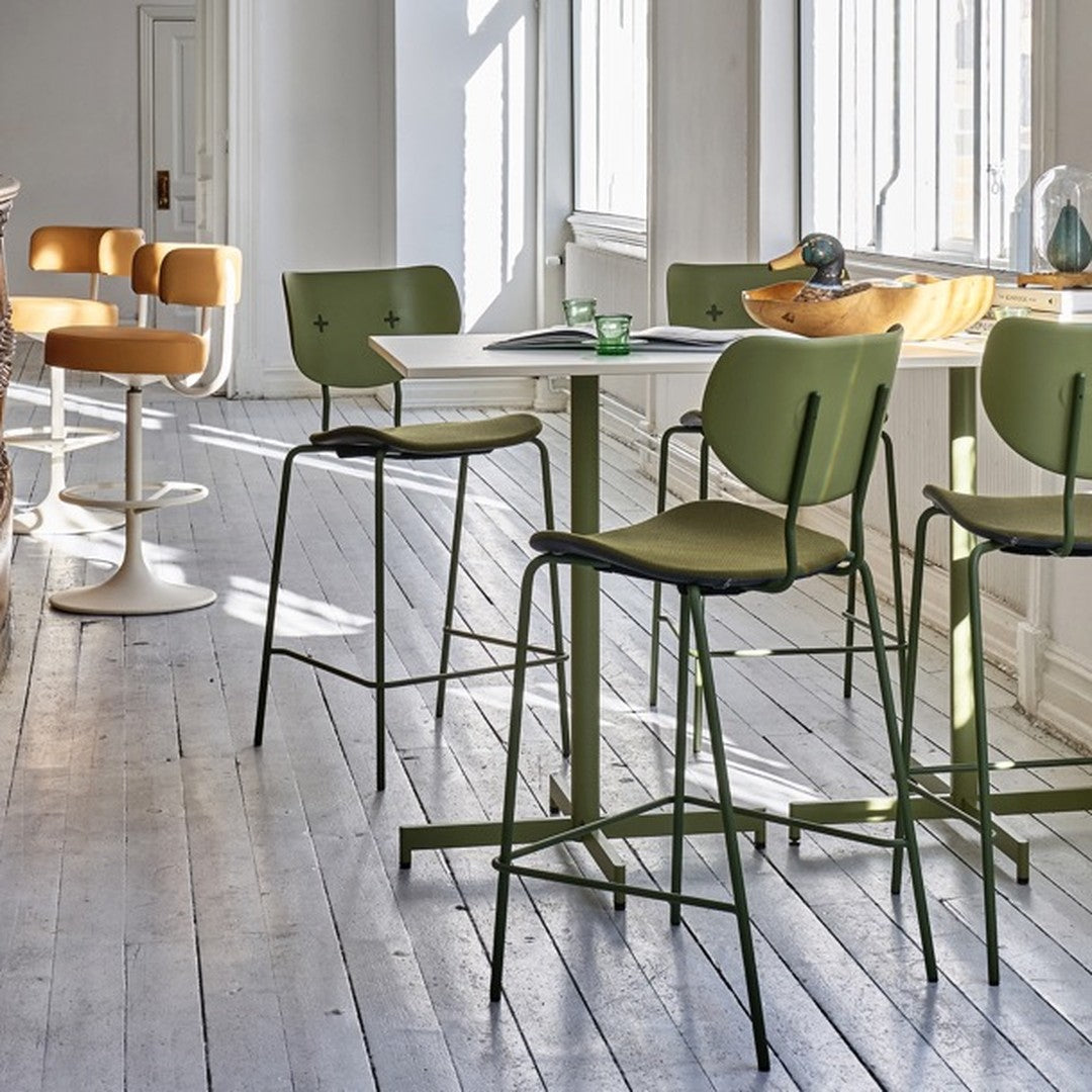 Modern green bar stools with warm tan accents, set against a light wooden floor and sunlit white walls.