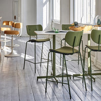 Modern green bar stools with warm tan accents, set against a light wooden floor and sunlit white walls.