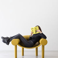 Yellow contemporary chair with rounded arms, paired with black apparel and a bright yellow book, set against a white background.