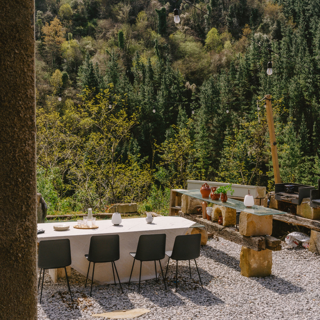 Outdoor dining setup featuring neutral furniture against a lush green forest backdrop and muted gravel patterns.