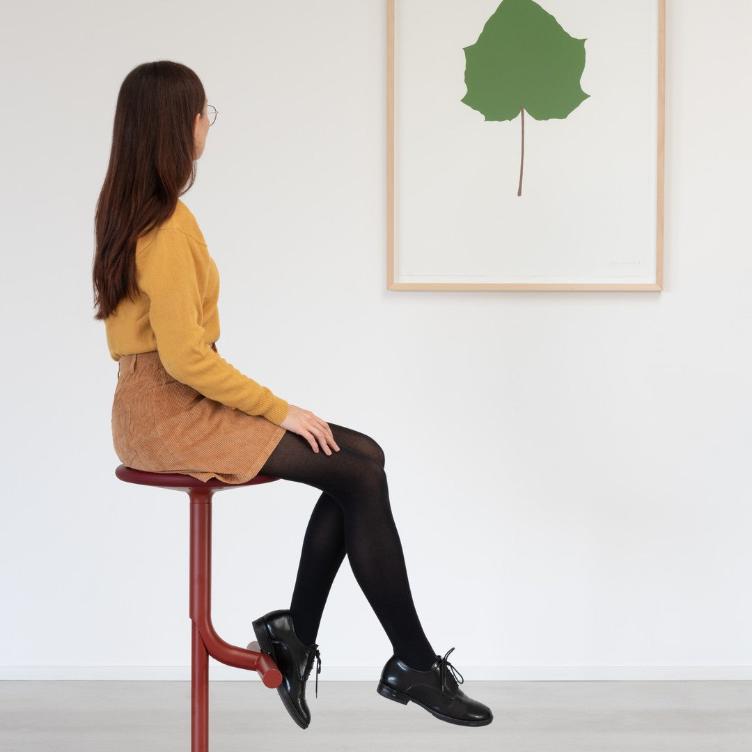 Woman in a mustard sweater, brown corduroy skirt, black tights, and black shoes; green leaf artwork in background.