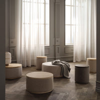 Neutral-toned round poufs with subtle ribbed patterns in a softly lit room with sheer white curtains.