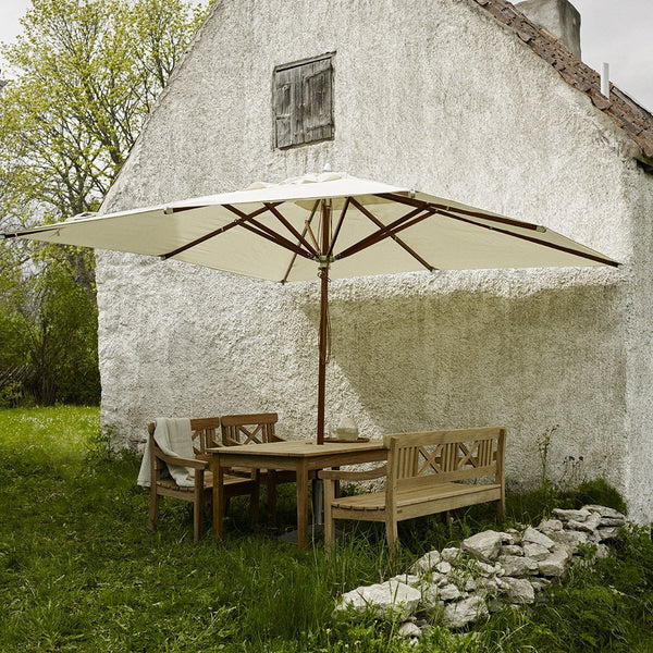 Cream-colored patio umbrella over a brown wood table and chairs in a lush green garden setting.
