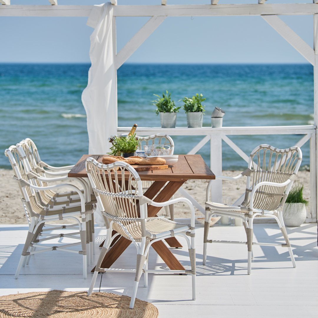 White wicker chairs and a brown wooden table on a beach porch, surrounded by greenery and blue ocean views.