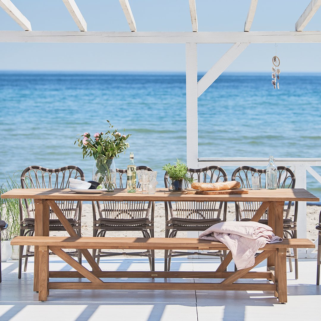 Stylish wooden dining table with beige and gray woven chairs by the beach, featuring a soft pastel throw.