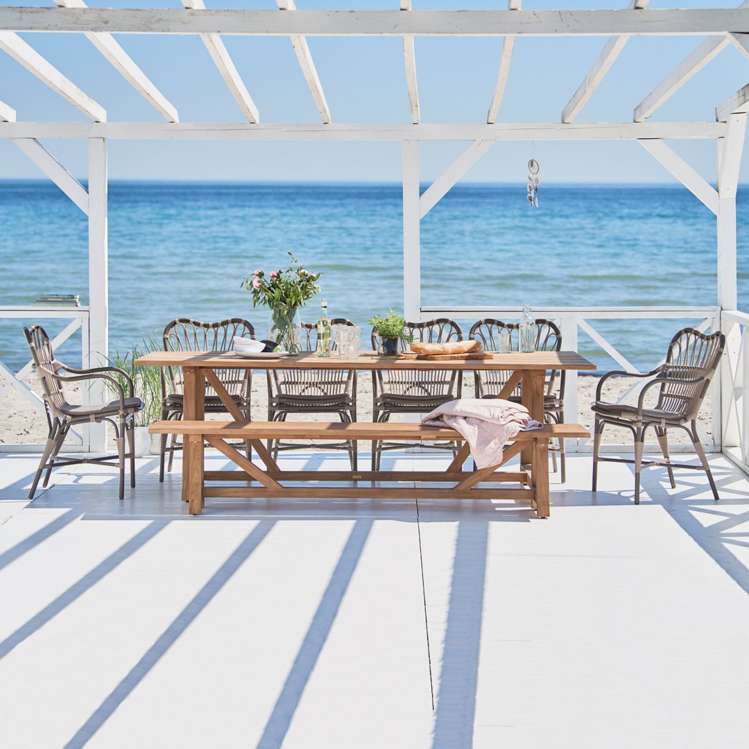 "Outdoor dining set with natural wood tones and white accents, featuring a coastal backdrop and striped shadows."